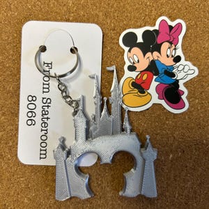以下が含まれることがあります： お城のデザインのシルバー色のキーホルダー。金属製のリングとチェーンが付いています。「From Stateroom 8066」と書かれた白いカードがキーホルダーの隣にあります。ミッキーとミニーマウスのステッカーも付いています。
