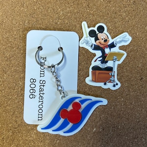 以下が含まれることがあります： 赤色のミッキーマウスの頭のロゴが白と青のデザインに施された、ディズニーのクルーズをテーマにしたキーホルダー。「From Stateroom 8066」の文字が入ったカードにキーホルダーが取り付けられています。指揮者の衣装を着たミッキーマウスのステッカーも付いています。
