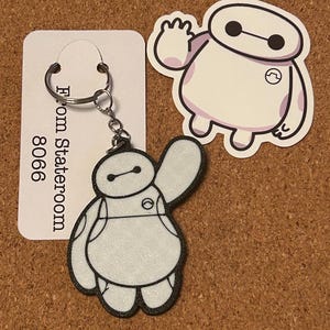 May include: A white and black Baymax keyring with a matching sticker. The keyring has a silver ring and chain. The sticker and keyring feature the character waving. A white tag with the text "From Stateroom 8066" is also present.