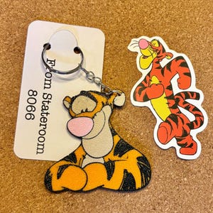 以下が含まれることがあります： ティガーのキーホルダーとステッカーのセット。キーホルダーは、オレンジ、黒、タン色のティガーのキャラクターが特徴です。ステッカーは、立っているポーズのティガーを示しています。「From Stateroom 8066」というテキストの白いタグも含まれています。
