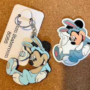 以下が含まれることがあります： ミッキーマウスのキーホルダーとステッカー。ミッキーは、ピンクのアクセントが付いた水色のバニーコスチュームを着ています。キーホルダーにはシルバーのリングと「From Stateroom 8066」と書かれたタグが付いています。ステッカーは、同じコスチュームを着たミッキーの別の画像です。