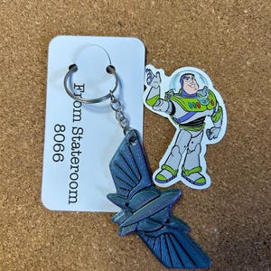 Op de afbeelding: Een sleutelhanger met een blauw en paars iriserend ruimteschipontwerp en een Buzz Lightyear-sticker. De sleutelhanger is bevestigd aan een wit label met de tekst "From Stateroom 8066".
