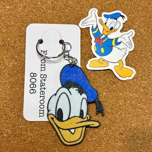 May include: A Donald Duck keyring with a blue hat and yellow beak, attached to a metal ring. A matching Donald Duck sticker is also present. The keyring and sticker are on a corkboard background.