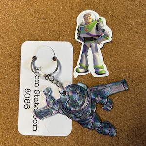 Op de afbeelding: Een sleutelhanger met een 3D-geprint Buzz Lightyear-ontwerp in paars, blauw en groen, met een zilveren sleutelring. Een Buzz Lightyear-sticker en een wit label met tekst zijn ook aanwezig.