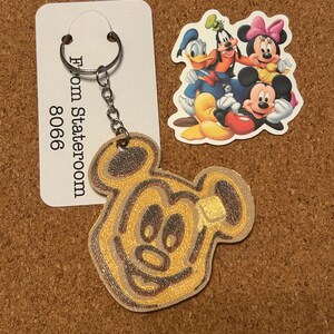 May include: A Mickey Mouse keyring with a gold and brown design, attached to a tag that reads "From Stateroom 8066." A sticker featuring Mickey Mouse, Minnie Mouse, Donald Duck, and Goofy is also present.
