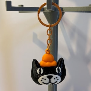 以下が含まれることがあります： オレンジ色の帽子をかぶった黒猫のキーホルダー。猫の目、鼻、口には白いディテールがあります。キーホルダーはオレンジ色のリングとチェーンに付いています。背景は明るいグレーです。