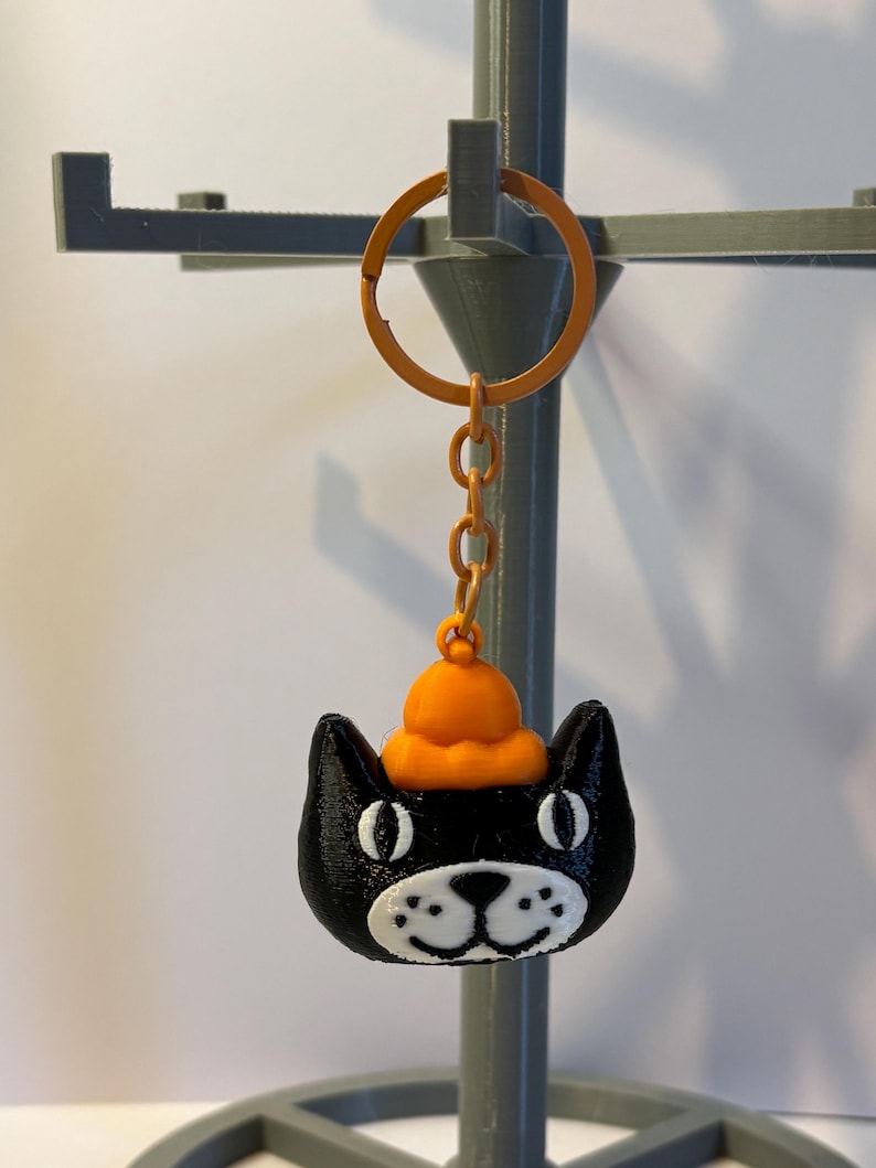 以下が含まれることがあります： オレンジ色の帽子をかぶった黒猫のキーホルダー。猫の目、鼻、口には白いディテールがあります。キーホルダーはオレンジ色のリングとチェーンに付いています。背景は明るいグレーです。