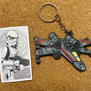 May include: A Star Wars keyring featuring a grey X-wing fighter with red and yellow detailing, attached to a metal ring and chain. A black and white card with a Stormtrooper illustration is also present.