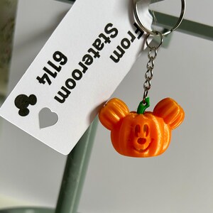 May include: Orange pumpkin-shaped keychain with Mickey Mouse ears and a carved face. The keychain is attached to a silver chain and a silver ring. A white tag with black text reads "Stateroom 6114".