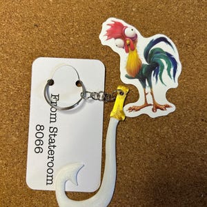 May include: A white fish hook keyring with a yellow handle. A cartoon rooster sticker with red, yellow, and blue colours is next to the keyring. A white tag with the text "From Stateroom 8066" is also present.