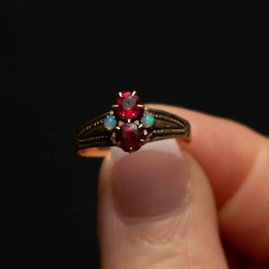 May include: A gold ring featuring three red gemstones and two smaller white gemstones. The ring is displayed against a black backdrop.