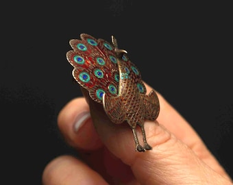 May include: A peacock-shaped brooch featuring a textured metallic body and a fanned tail decorated with red enamel and small blue circular accents. The piece displays intricate metalwork details across the wings and tail feathers in a compact design.