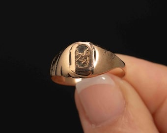 Könnte beinhalten: Ein goldener Siegelring mit einem erhabenen Design auf der Oberseite. Der Ring wird an einem Finger getragen.