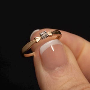 May include: A gold ring with a small diamond in the centre. The ring is designed with a bow shape.