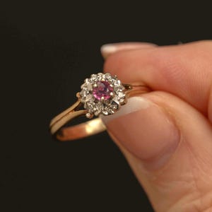 May include: A gold ring with a central pink gemstone and surrounding white stones. The ring has a delicate band and a halo design. The ring is set against a dark background.