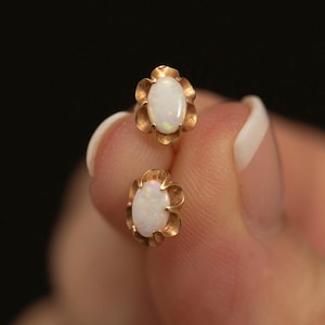 May include: A pair of stud earrings featuring oval-shaped white opals set in gold-toned metal. Each opal is surrounded by a scalloped, petal-like metal frame that creates a flower-inspired silhouette. The earrings have a polished, metallic finish.