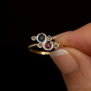 Könnte beinhalten: Ein Goldring mit einer Ansammlung kreisförmiger Fassungen, die einen zentralen blauen Edelstein, einen zentralen rosa Edelstein und vier kleinere klare Akzentsteine in einem versetzten, asymmetrischen Muster auf einem dünnen Goldband enthalten.