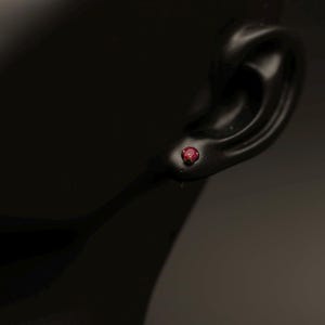 May include: A small, round, deep red gemstone stud earring secured in a metal setting. The earring is positioned in the earlobe of a dark, smooth, mannequin-style display form, highlighting the vibrant colour and circular shape of the stone.