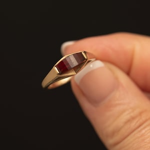 May include: A gold ring featuring a rectangular, deep red gemstone set horizontally within a smooth, polished metal band. The setting has a geometric, tapered design that frames the dark red stone, creating a clean and structured appearance.