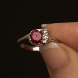 May include: A silver ring with a large, round, deep red gemstone and a cluster of smaller, clear stones. The ring is held against a black background, highlighting the jewellery's design and sparkle.