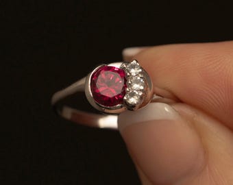 May include: A silver ring featuring a large, round, deep red gemstone and a cluster of smaller, clear stones. The ring is held against a black background, showcasing the jewelry's design and sparkle.