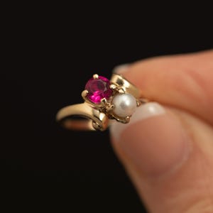 May include: A gold bypass ring featuring a round, faceted deep pink gemstone and a small, round white pearl. The two stones are set side-by-side on a polished gold band that curves around the finger, creating a classic and elegant jewellery piece.