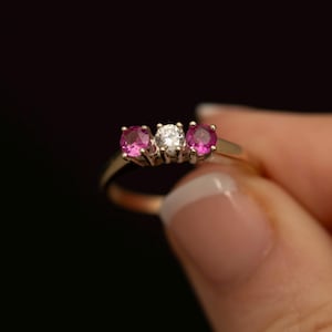 May include: A gold ring featuring a three-stone setting with a central clear round-cut gemstone flanked by two vibrant pink round-cut gemstones. The stones are held in place by metal prongs on a polished gold band against a dark background.