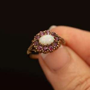 Könnte beinhalten: Ein Goldring mit einem zentralen, ovalen weißen Opal, der von einem Kranz aus zwölf kleinen, runden, tief rosa-roten Edelsteinen umgeben ist. Die Metallschiene weist an den Seiten ein dekoratives, strukturiertes Design auf.
