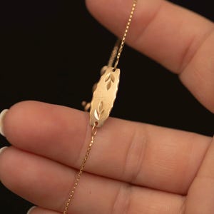 Könnte beinhalten: Ein goldfarbenes Armband mit einer zentralen, rechteckigen Metallplatte mit gewellten Kanten und zwei eingravierten Blattmotiven. Die Platte ist an einer zarten, dünnen goldfarbenen Kette befestigt, die über die Finger einer Hand verläuft.