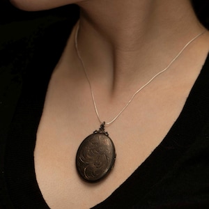 May include: An oval-shaped dark metal locket featuring an intricate engraved floral pattern on its surface. The locket is suspended from a thin, delicate silver-toned chain that rests against the neck, providing a classic and understated appearance.