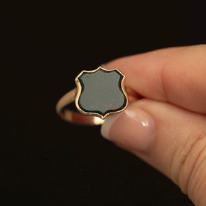 Peut inclure: Une bague en or dotée d'un chaton en forme de blason avec une incrustation de pierre noire lisse. L'anneau en métal est poli et reflète la lumière, tandis que la pierre centrale présente une surface plane avec de légères taches brun-rouge.