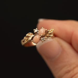 May include: A gold ring featuring two oval-shaped, light pink cabochon stones positioned vertically in the center. The stones are flanked by two small, clear, round-cut stones and decorative gold leaf-shaped metalwork on both sides of the band.