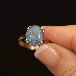 May include: A gold ring featuring a large, oval-shaped cabochon opal gemstone. The opal displays a play of colour with flecks of blue, green, and orange against a dark, translucent base. The stone is secured by six small gold prongs on a band.