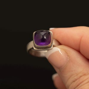 Könnte beinhalten: Ein Silberring mit einem glatten, kissenförmig geschliffenen violetten Amethyst-Edelstein, der in einer einfachen Metallfassung sitzt. Die Ringschiene besteht aus poliertem Silber und hat eine schlichte, massive Form, die den quadratischen Stein hält.