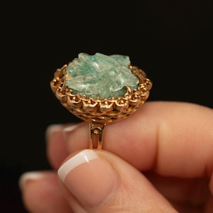 May include: A gold-toned ring featuring a cluster of light green, raw, crystalline stones set within a decorative, scalloped metal frame. The setting has an intricate, openwork design around the edges that holds the textured, pale green mineral cluster securely in place on a simple, thin metal band.