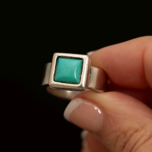 May include: A silver ring featuring a square-shaped, smooth turquoise stone set in a simple metal bezel. The band is plain and polished, showcasing a clean design with the vibrant blue-green gemstone as the central focal point of the piece.
