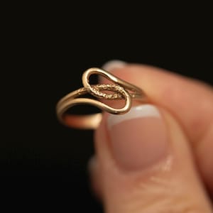 May include: A gold ring featuring a continuous, fluid band that loops over itself to create an open, asymmetrical shape. The central portion of the metal band displays a textured, granular surface finish, while the outer edges remain smooth.