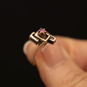 May include: A gold ring featuring a small, round, vibrant pink gemstone set in a prong setting. The band is constructed with a geometric, open-work design consisting of interconnected rectangular metal bars that frame the central stone.