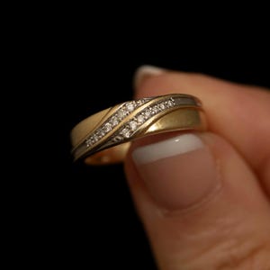 Könnte beinhalten: Ein zweifarbiger Ring mit einem gebürsteten Gelbgoldband und einer diagonalen Vertiefung, die mit einer Reihe kleiner, klarer, funkelnder Steine besetzt ist. Das Band hat einen polierten silberfarbenen Akzent parallel zu den Steinen.