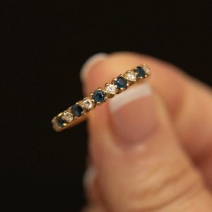 以下が含まれることがあります： 丸くカットされた濃い青色のサファイアと透明な白いダイヤモンドが交互に並んだゴールドの指輪。細く磨かれたイエローゴールドのバンドの上半分に、石が途切れることなく一列にセットされています。