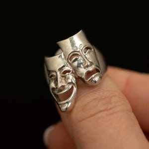 Peut inclure: Une bague en argent représentant les masques de théâtre comique et tragique. La bague montre un visage souriant et un visage triste, symbolisant la dualité des émotions. La bague est présentée sur un fond noir.