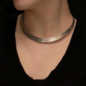Könnte beinhalten: Eine silberfarbene Metall-Halskette in Choker-Form mit einem breiten, geschwungenen Band und einer gehämmerten, strukturierten Oberfläche. Das Schmuckstück liegt flach am Hals an und zeigt eine reflektierende Fläche mit unregelmäßigen Vertiefungen.
