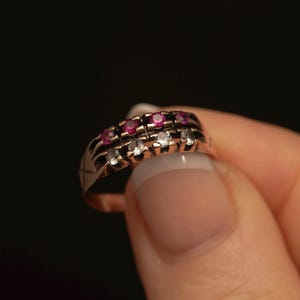 May include: A gold ring with two rows of gemstones. The top row features small, round, red gemstones, while the bottom row has clear, white gemstones. The ring is held against a black background.