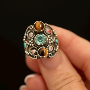 Peut inclure: Une bague en argent ornée d'une pierre centrale bleu clair à facettes, entourée de deux cabochons en œil-de-tigre marron. La monture métallique présente des motifs complexes en fil torsadé et de petits accents circulaires rouges et turquoise.