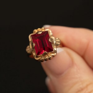Könnte beinhalten: Ein goldener Ring mit einem rechteckigen, facettierten tiefroten Edelstein, der in eine dekorative Metallfassung mit gewellten Rändern eingelassen ist. Die Fassung weist ein strukturiertes, verziertes Design auf, das den zentralen Stein umgibt.