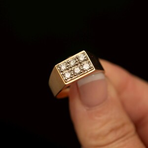 May include: A gold ring featuring a rectangular head set with six small, clear, round-cut diamonds arranged in two rows of three. The band is smooth and polished, showcasing a solid gold construction that tapers slightly towards the back of the ring.