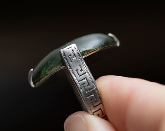 May include: A silver ring with a green stone set in a bezel setting. The band of the ring is decorated with a Greek key pattern.