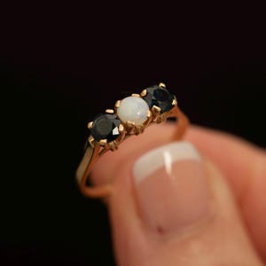 May include: A gold ring featuring a central round white opal flanked by two round black gemstones. The stones are set in a prong setting on a thin gold band. The design showcases a three-stone arrangement with a polished metal finish.