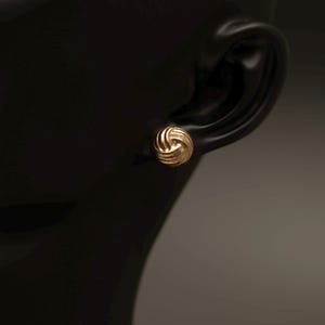 Peut inclure: Une boucle d'oreille à clou en or présentant une forme sphérique avec un motif de nœud texturé et tourbillonnant. La boucle d'oreille est polie et reflète la lumière, montrant une série de lignes arrondies interconnectées convergeant vers le centre.