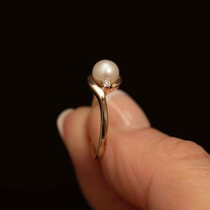 May include: A gold ring featuring a white pearl and a small diamond accent. The ring has a delicate, curved design, with the pearl set at the top. The ring is held against a dark background.